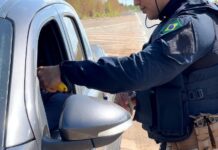 PRF prende três motoristas por embriaguez, autua 20 e recupera moto furtada na BR-174 Policial da PRF de uniforme escuro e colete tático aborda o motorista de um carro prata em uma rodovia, segurando um etilômetro amarelo na janela do veículo.