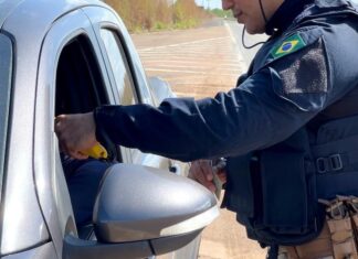 PRF prende três motoristas por embriaguez, autua 20 e recupera moto furtada na BR-174 Policial da PRF de uniforme escuro e colete tático aborda o motorista de um carro prata em uma rodovia, segurando um etilômetro amarelo na janela do veículo.