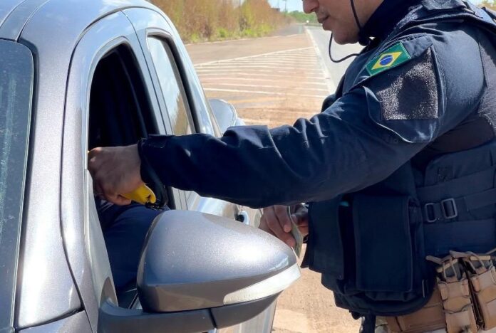 Policial da PRF de uniforme escuro e colete tático aborda o motorista de um carro prata em uma rodovia, segurando um etilômetro amarelo na janela do veículo.